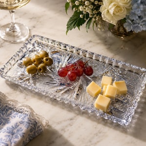May include: A rectangular clear glass serving tray featuring a faceted starburst pattern and a scalloped edge. The tray holds a small cluster of green olives, a bunch of red grapes, and several cubed pieces of yellow cheese arranged on its surface.