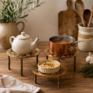 May include: A kitchen scene featuring a white teapot, a copper saucepan with soup, and a small dish of food, all on brass trivets. A beige jug and wooden spoons are in the background, on a wooden surface.