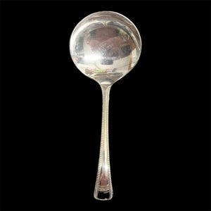 May include: A silver soup ladle with a round bowl and a long handle. The handle has a beaded design along the edges and a decorative shape at the end. The ladle is set against a black background.