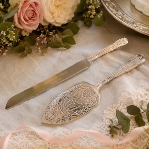 May include: Silver cake knife and server set with intricate floral patterns. The knife blade is engraved with the names "Andrew and David" and the date "09-09-2007". The set is presented on a neutral surface with floral decorations.