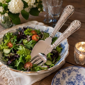 May include: A large, scalloped-edge ceramic bowl with a blue and white floral pattern filled with a fresh green salad containing sliced cucumbers and cherry tomatoes, accompanied by two ornate silver-plated serving utensils with shell motifs.
