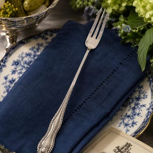 May include: A silver-plated pickle fork featuring a long, slender handle with an ornate, embossed border and a three-pronged head. The handle terminates in a decorative oval motif, showcasing intricate metalwork and a polished, reflective finish.