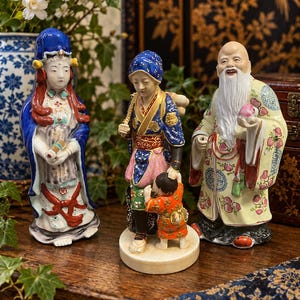 May include: Three porcelain figurines featuring a standing figure in a blue robe, a figure carrying a child in a blue and pink patterned outfit, and an elderly figure with a long white beard holding a peach, all with intricate painted details.