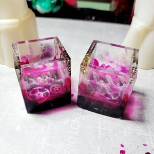 Puede incluir: Dos pequeñas cajas de vidrio transparente con detalles dorados. Las cajas están llenas de resina rosa y blanca y contienen pequeños engranajes. Las cajas están sobre una superficie blanca.