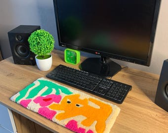 May include: A colorful, abstract rug with a yellow, orange, pink, and green design. The rug is on a wooden desk with a computer monitor, keyboard, and two speakers.