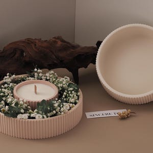 Puede incluir: Un portavelas de cerámica rosa con una vela blanca y un anillo de flores blancas alrededor de la vela. El portavelas tiene una textura acanalada.