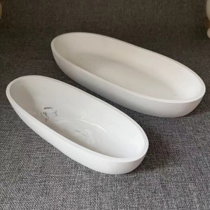 Könnte beinhalten: Zwei weiße ovale Keramikschalen, eine kleiner als die andere, auf einer grauen strukturierten Oberfläche.