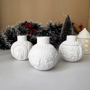 Puede incluir: Tres portavelas de cerámica blanca con forma de bolas de Navidad con diferentes diseños. Las bolas presentan diseños en relieve de un reno, una escena de la Natividad y un ángel de Navidad.