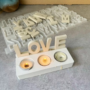 May include: White wooden candle holder with the word "LOVE" spelled out in large letters. The holder has three candle wells with lit tea lights.