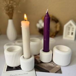Puede incluir: Portavelas de cerámica blanca con un diseño acanalado. Dos velas están en los portavelas, una blanca y una morada.