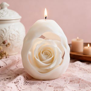 Può includere: Una candela a forma di rosa color crema con uno strato esterno scolpito. La candela è accesa e posizionata su una tovaglia di pizzo rosa. Altre candele e un vaso di ceramica bianco sono sullo sfondo.