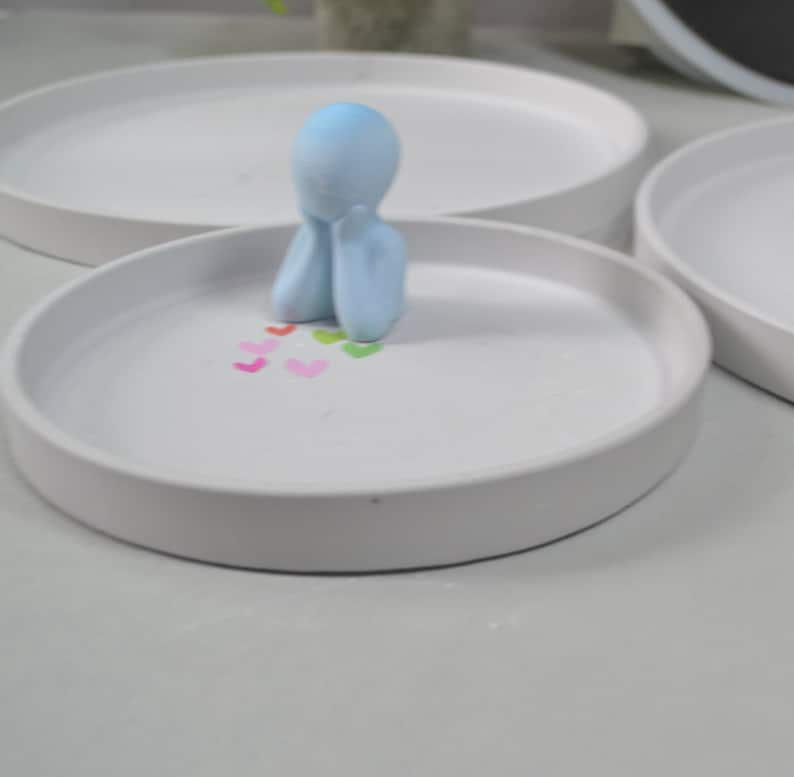 Pu&ograve; includere: Un piatto in ceramica bianca con una statuetta azzurra di una persona seduta con la testa tra le mani. Il piatto &egrave; decorato con piccoli cuori colorati.