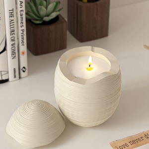 Può includere: Una candela bianca accesa in un portacandele a forma di uovo strutturato, con un coperchio abbinato. La candela è su una superficie bianca, con libri e una piccola pianta sullo sfondo. La fiamma della candela è gialla.