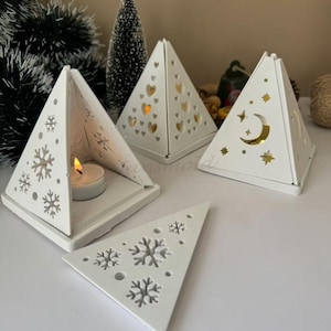 Può includere: Tre portacandele in legno bianco a forma di piramide con diversi disegni ritagliati. I disegni includono fiocchi di neve, cuori e una mezzaluna e stelle. I portacandele sono su una superficie bianca.