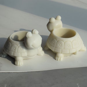 Puede incluir: Dos macetas de cerámica blanca en forma de tortuga con una pequeña abertura en la parte superior para plantar. Las tortues tienen un diseño de caparazón detallado y una cara sonriente.