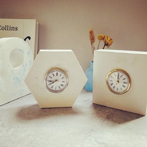 May include: Three white marble clocks with gold hands and Roman numeral faces. The clocks are in different shapes, including a hexagon and a rectangle.
