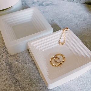 May include: Two white ceramic trays with a layered design. The trays are square and have a raised edge. The smaller tray is on top of the larger tray. There are gold earrings and a gold necklace in the smaller tray.