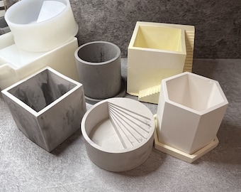 May include: A set of six different concrete planter molds. The molds are in various shapes, including square, round, and hexagonal. The molds are made of a light gray concrete and have a smooth finish.