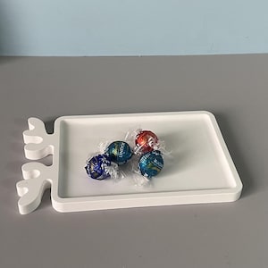 May include: A white rectangular tray with a decorative antler-like design on one side. The tray holds four wrapped chocolate candies in blue and red foil. The background is a light blue and gray.