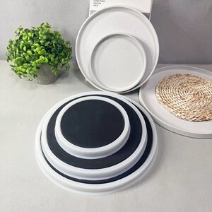 Pu&ograve; includere: Set di quattro sottobicchieri rotondi bianchi con un centro nero. I sottobicchieri sono impilati uno sopra l'altro. I sottobicchieri sono su una superficie bianca.