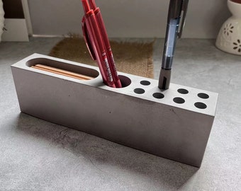 May include: A gray concrete desk organizer with multiple compartments for pens and pencils. Three red pens and one black pen are in the organizer.