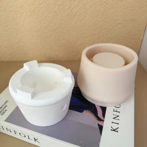 以下が含まれることがあります： コンクリート製のキャンドルホルダーを作るための白いシリコンモールド2つ。モールドは、「KINFOLK」と印刷された本の表紙の上に置かれています。