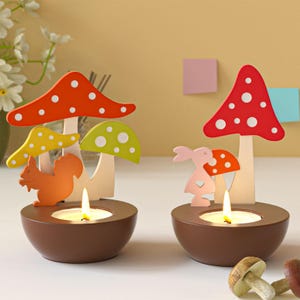 Op de afbeelding: Twee decoratieve kandelaars met paddenstoel- en bosdierontwerpen. Elke houder heeft een bruine komvormige basis met een brandende kaars, bekroond met kleurrijke houten paddenstoelen en een eekhoorn of konijn. De paddenstoelen hebben rode, oranje en gele doppen.