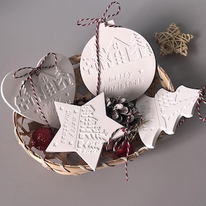 Pode incluir: Enfeites de Natal brancos em forma de coração, estrela, círculo e árvore, com a inscrição "merry Christmas". Estão numa cesta de vime com uma pinha e enfeites vermelhos.
