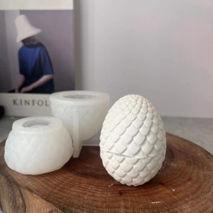 Op de afbeelding: Drie witte siliconen mallen voor het maken van kaarsen. De mallen zijn gevormd als een dennenappel, een kleine kom en een drakenei.