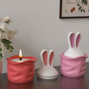Könnte beinhalten: Eine brennende rote Kerze in einem Keramikbehälter, neben einem weißen hasenförmigen Behälter mit rosa Innenohren und einem rosa Behälter mit hasenförmigem Deckel. Die Gegenstände stehen auf einer dunklen Oberfläche, mit einem Blumenarrangement und gerahmten Kunstwerken im Hintergrund.