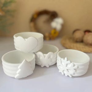 以下が含まれることがあります： 異なるホリデーテーマのデザインが施された白いセラミックボウル4個。ボウルには、ハート、雪片、クリスマスツリー、蝶が描かれています。