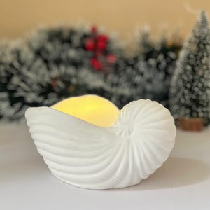 Op de afbeelding: Witte keramische schelp kaarsenhouder met een warm wit LED-licht erin. De schelp is getextureerd en heeft een spiraalvormig ontwerp.