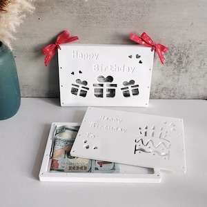 Może przedstawiać: Białe pudełko z kartonu z wyciętym wzorem prezentów i tekstem "Happy Birthday". Pudełko jest otwarte, aby ujawnić mniejsze białe pudełko z kartonu z wyciętym wzorem tortu urodzinowego i tekstem "Happy Birthday To:".