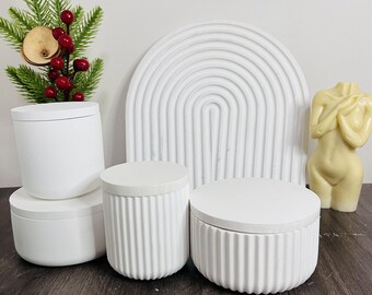 Può includere: Quattro contenitori in ceramica bianca con coperchi. I contenitori hanno un design a coste e sono impilati uno sopra l'altro. I contenitori sono su una superficie di legno.