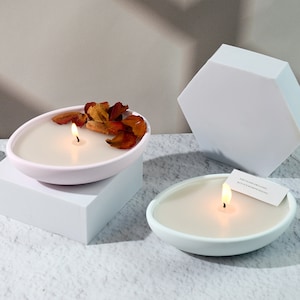 Może przedstawiać: Dwie owalne świece z zapalonymi knotami. Jedna świeca jest w różowym naczyniu i ozdobiona suszonymi płatkami. Druga świeca jest w białym naczyniu z małą kartką z napisem "THE YEARS ARE LONG BUT IT'S WORTH SEEING".