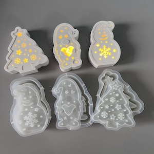 Op de afbeelding: Een verzameling kerst-thema hars mallen en afgewerkte lichtgevende decoraties. De mallen omvatten een sneeuwpop, een gnome en een kerstboom. De afgewerkte decoraties zijn wit met gele binnenverlichting. De gnome heeft een hartvormig licht.