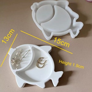 Puede incluir: Molde de silicona blanco con forma de pez con dos compartimentos. El molde mide 13 cm de largo, 15 cm de ancho y 1,9 cm de alto.