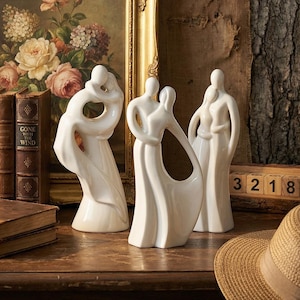 May include: Three white ceramic sculptures depicting embracing couples. The sculptures have a minimalist design with smooth, flowing lines. The figures are in various poses of affection, with one sculpture featuring an open design. The sculptures are displayed on a wooden surface.