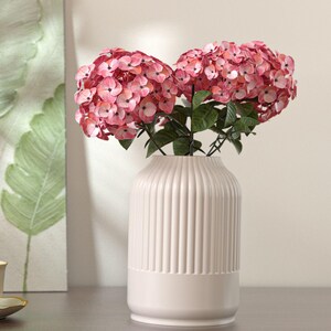 Può includere: Un vaso in ceramica bianco con dettagli a coste verticali, che contiene un bouquet di ortensie rosa e rosse con foglie verdi. Il vaso ha una base arrotondata e una sezione superiore cilindrica.