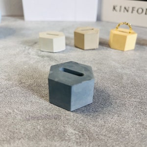Può includere: Tre porta anelli esagonali in cemento. I porta anelli sono di diversi colori: bianco, beige e grigio. Il porta anelli grigio è a fuoco.