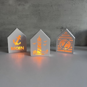 Puede incluir: Tres luces decorativas blancas con forma de casa y temas náuticos. Una presenta un ancla y la palabra "MOIN", otra un faro y la tercera un ancla dentro de un círculo. Cada luz emite un brillo cálido y anaranjado.