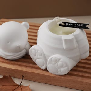 Pode incluir: Dois itens brancos em forma de urso estão em uma superfície de madeira. Um é uma pequena estatueta de urso, e o outro é uma vela em forma de urso com um pavio e uma etiqueta que diz "HANDMADE Especially for You."