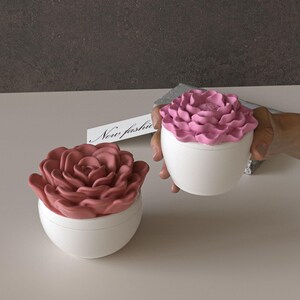 Puede incluir: Dos recipientes de cerámica blanca con tapas en forma de rosa. Uno tiene una tapa rosa polvorienta y el otro una tapa rosa. El texto "New fashion" está en una pequeña tarjeta blanca. Los recipientes están sobre una superficie blanca.