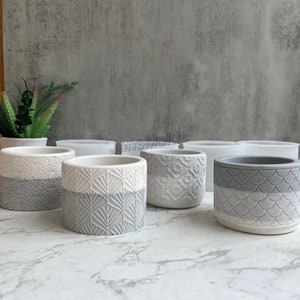 May include: A set of eight gray concrete planter molds with different geometric patterns. The molds are arranged in two rows of four. The molds are made of silicone and are designed to create unique and stylish planters.
