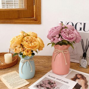 May include: Two ceramic vases, one light blue and one pink, each holding a bouquet of flowers. The blue vase has yellow roses, and the pink vase has pink roses. Both vases are tied with twine. The scene is set on a wooden surface.