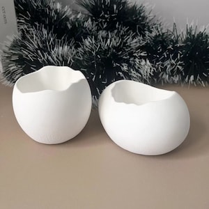 Pode incluir: Dois vasos de cerâmica branca em forma de ovo com uma rachadura no topo. Os vasos estão sobre uma superfície marrom.