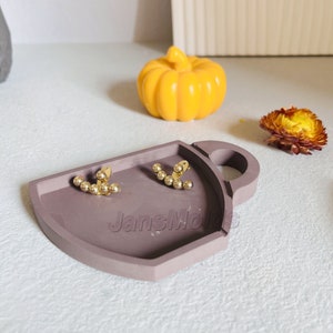 Puede incluir: Un par de pendientes de tono dorado con un diseño curvo que presenta una fila de pequeñas cuentas redondas. Los pendientes se muestran en una bandeja gris texturizada con un asa.