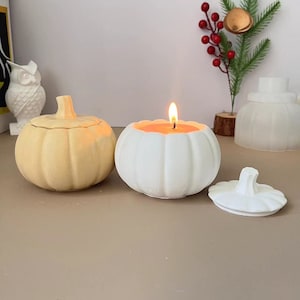 Puede incluir: Dos portavelas con forma de calabaza, uno blanco y otro beige. El portavelas blanco tiene una vela encendida en su interior. El portavelas beige está cerrado.