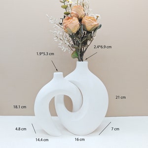 Puede incluir: Dos jarrones de cerámica blanca, uno con un diseño curvo y abstracto, el otro con una forma redonda. Los jarrones están conectados, creando un aspecto único y moderno. El jarrón más grande mide 21 cm de alto y el jarrón más pequeño mide 18,1 cm de alto.