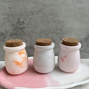 Puede incluir: Tres pequeños frascos decorativos con tapas de corcho. Los frascos están hechos de cerámica blanca con un diseño de mármol. Los frascos están en una bandeja rosa y blanca.